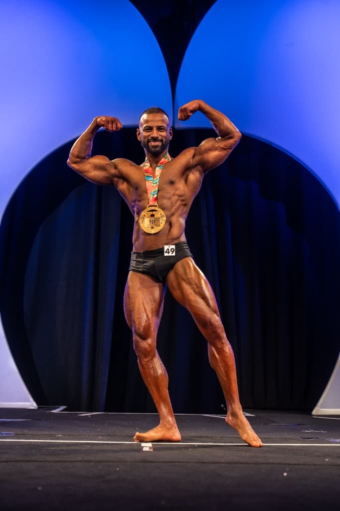 Puneet Sharma on the competition stage in a front double biceps pose, wearing a 1st place gold medal